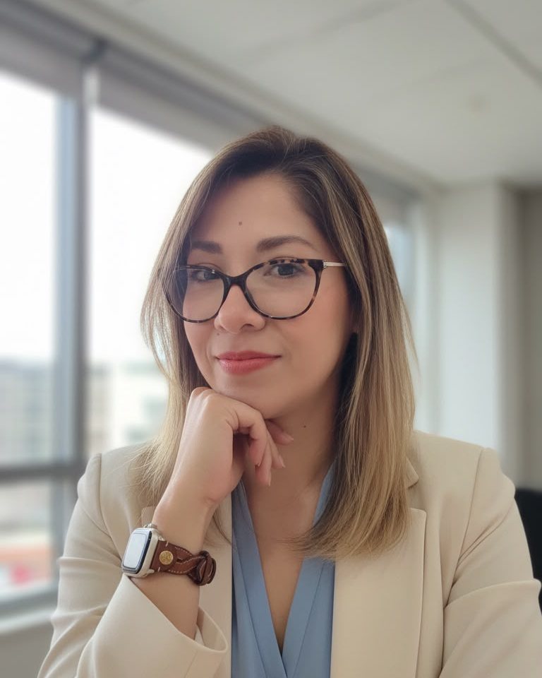 La Dra. Lucy Tania Ruiz Segura en su consultorio, con lentes y saco claro, la barbilla apoyada en la mano.